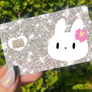 May include: Glittery silver card with a white bunny graphic and a pink flower. The card has a heart-shaped cutout with a chip. The card is held in a hand with a manicured nail.