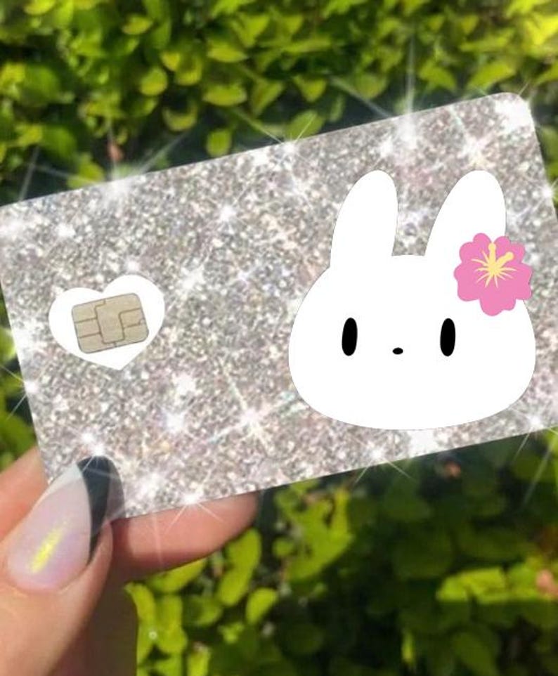May include: Glittery silver card with a white bunny graphic and a pink flower. The card has a heart-shaped cutout with a chip. The card is held in a hand with a manicured nail.