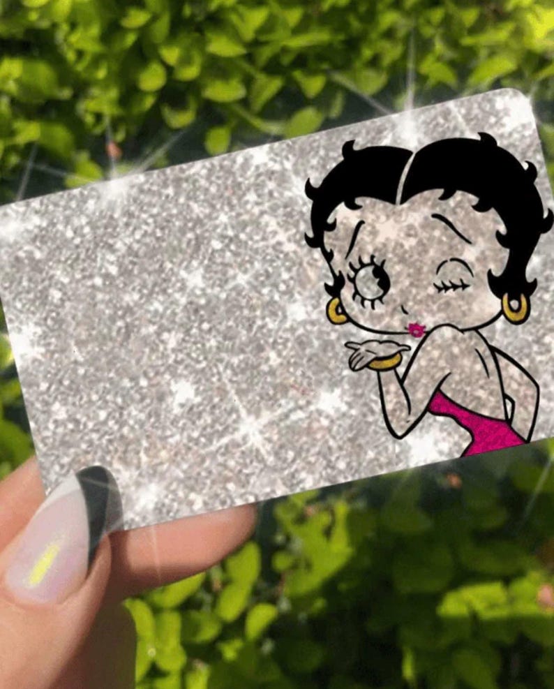 May include: A silver glitter card featuring a cartoon image of Betty Boop blowing a kiss. The card has a rectangular shape and is held by a hand with a manicured nail. The background is a blurred green.