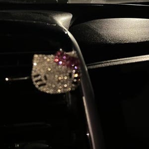 Op de afbeelding: Een Hello Kitty-vormige auto-luchtverfrisser clip bedekt met sprankelende zilveren strass-steentjes met een roze strass-strik, bevestigd aan een donker ventilatierooster van het dashboard in een omgeving met weinig licht en een metallic afwerking.