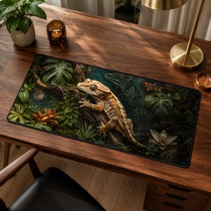 May include: A rectangular desk mat featuring a detailed illustration of a crested gecko perched on a textured tree branch. The design includes lush green monstera leaves, ferns, orange bromeliads, and white flowers against a dark forest background.