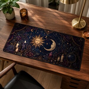 Può includere: Un tappetino da scrivania rettangolare con un design celeste blu scuro e viola, caratterizzato da un sole e una luna con volto centrale, circondati da illustrazioni di cristalli, candele accese, tralci di fiori e un globo stellato.