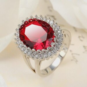 Puede incluir: Anillo de plata con una gran gema roja brillante de corte ovalado, rodeada por un halo de gemas claras más pequeñas. La banda del anillo es de plata pulida, realzando el brillo de la piedra central.