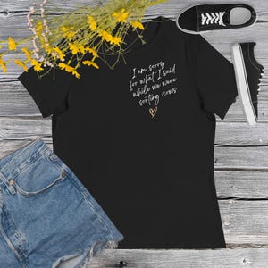 May include: Black t-shirt with the text "I am sorry for what I said while we were sorting cows" in white script, with a small heart. The shirt is paired with denim shorts and black sneakers.