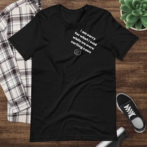 May include: Black t-shirt with white text that reads "I am sorry for what I said while we were sorting cows" and a smiley face. The shirt is displayed on a wooden surface with a plaid shirt, a succulent plant, and a pair of black sneakers.