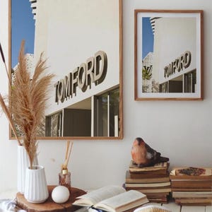May include: Two framed prints with the TOM FORD logo, a decorative bird figurine, books, and a cup of coffee on a white table. A vase with dried pampas grass and a straw hat complete the decor.