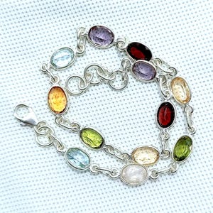 May include: A silver bracelet with a series of oval gemstones in colours such as purple, red, yellow, green, and blue. The bracelet has a lobster clasp and is displayed on a white surface.