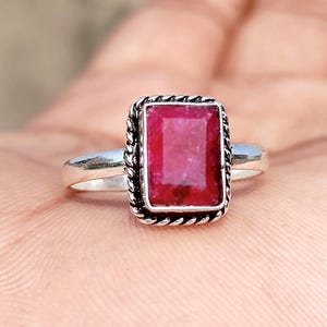 May include: A silver ring featuring a rectangular, faceted deep red gemstone set in a twisted rope-style metal border. The band is smooth and polished, holding the central stone securely in a bezel setting with a distinct textured edge detail.