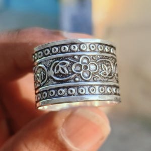 May include: A silver-colored ring with intricate floral and leaf patterns. The ring features a series of small, circular designs along the top and bottom edges, with detailed embossed floral and leaf motifs in the center.