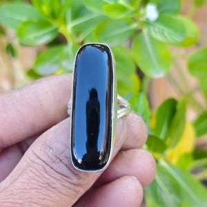 May include: A ring featuring a long, vertical, rectangular black stone set in a polished silver-toned metal band. The stone has a smooth, reflective surface and is securely held by a simple metal bezel setting on a plain, slender band.
