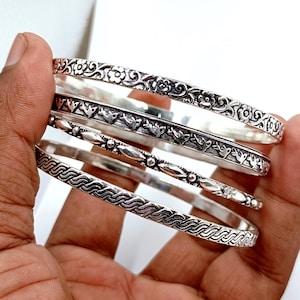 Könnte beinhalten: Eine Sammlung silberner Armreifen mit aufwendigen Designs. Die Armreifen weisen florale, Vogel- und geometrische Muster auf. Die Armbänder werden in einer Hand gehalten, was die detaillierte Handwerkskunst und die unterschiedlichen Texturen jedes Stücks zeigt.