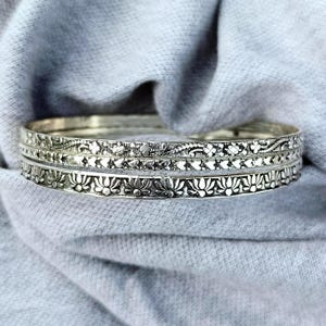 May include: Three silver bangles with intricate floral and leaf patterns. The bangles are stacked, showcasing detailed designs of flowers, leaves, and other decorative elements. The jewelry is set against a soft, gray fabric background.