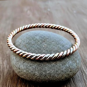 May include: A twisted metal bracelet with a mix of gold, silver, and copper tones. The bracelet is resting on a gray stone, with a wooden surface in the background. The bracelet's design features a rope-like texture.