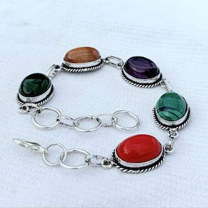 Pode incluir: Uma pulseira de prata com pedras preciosas ovais em várias cores: verde, castanho, roxo, vermelho e verde. As pedras são engastadas numa moldura de prata com uma borda em forma de corda. A pulseira tem cerca de 18 cm de comprimento.