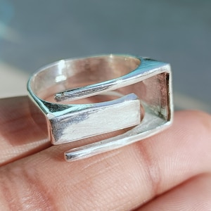 May include: A silver ring with a modern, geometric design. The ring features a rectangular centerpiece and a unique open band. The metal has a polished finish, reflecting light. This piece of jewelry is suitable for everyday wear.