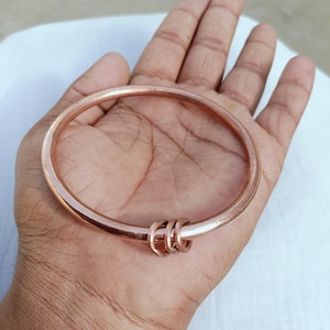 May include: A smooth, circular copper-toned bangle bracelet featuring a polished metallic finish. The bangle is designed with a single, continuous band that includes three small, matching copper-toned rings threaded onto the main structure.