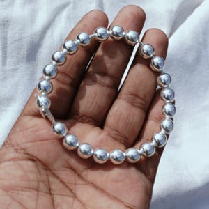May include: A silver bracelet featuring a series of round, polished beads strung together. The bracelet is shown on a hand, with the beads reflecting light. The clasp is visible, indicating how the bracelet is secured.