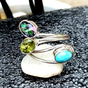 Może przedstawiać: Srebrny pierścionek z trzema owalnymi kamieniami szlachetnymi: zielono-fioletowym iryzującym kamieniem, jasnozielonym kamieniem i turkusem. Pierścionek leży na białym kamieniu na tle czarnego tła.