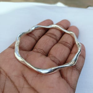 May include: A polished silver bangle bracelet featuring a continuous, undulating, wavy design. The metal surface is smooth and reflective, creating a series of gentle curves around the entire circumference of the circular jewelry piece.