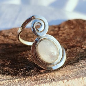 May include: A silver ring with a large, oval, light-colored stone. The ring features a spiral design above the stone. The ring is set on a brown surface.