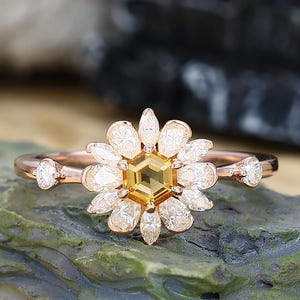 May include: A rose gold ring featuring a daisy design. The center stone is a yellow hexagon, surrounded by white petal-shaped stones. Additional white stones are set on the band on either side of the daisy. The ring is displayed on a green rock.