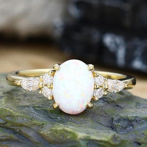 Può includere: Un anello d'oro con un grande opale bianco ovale come pietra centrale. L'anello è impreziosito da pietre preziose più piccole e trasparenti e montato su una fascia d'oro. L'anello è esposto su una superficie strutturata verde-grigia.