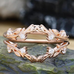 Puede incluir: Protector de anillo de oro rosa con un diseño de hojas y vides. El anillo presenta pequeñas piedras preciosas claras engastadas en las hojas. El anillo se muestra sobre una superficie de piedra verde texturizada.