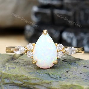 May include: A gold ring featuring a pear-shaped opal center stone, flanked by two small round diamonds and one marquise-cut diamond on each side. The band is thin and polished, showcasing a prong setting that secures the iridescent opal gemstone.