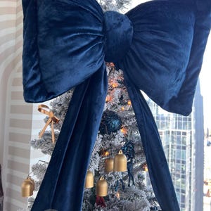 May include: A large, deep navy blue velvet bow tree topper with two long, flowing fabric tails. The bow is positioned at the top of a flocked Christmas tree decorated with small gold bells, dark blue ornaments, and gold ribbon accents.