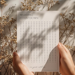 Puede incluir: Un calendario de cuenta regresiva de boda con una cuadrícula de números, sostenido por manos. La parte superior dice "WEDDING COUNTDOWN". La parte inferior dice "one year until".