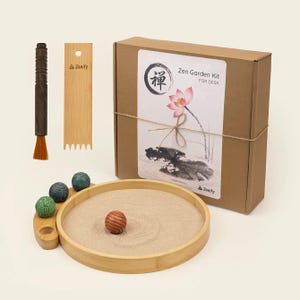 Può includere: Un kit da giardino zen per scrivania in una scatola di cartone marrone con un'illustrazione di fiore di loto e il testo Zen Garden Kit for Desk. Include un vassoio rotondo in bambù con sabbia, un rastrello in legno, una spazzola e quattro sfere testurizzate.