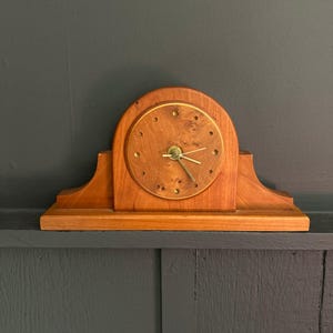 May include: A wooden mantel clock with a light brown finish. The clock has a round face with gold-colored hands and hour markers. The clock is approximately 10 inches wide and 7 inches tall.