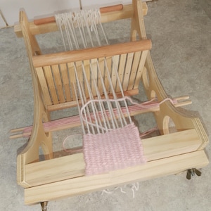 May include: A wooden weaving loom with white and pink yarn. The loom is made of light-coloured wood and features a partially woven pink textile. The loom is designed for creating small woven items.
