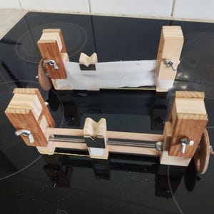 May include: Two wooden craft clamps with light and dark wood elements. Each clamp features a central support and adjustable screws. A piece of white fabric is held between the clamps.
