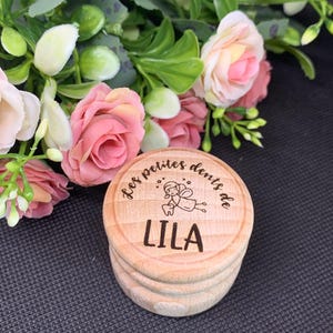 Könnte beinhalten: Eine kleine, runde Holzkiste mit dem Text "Les petites dents de LILA" und einer Feenillustration, die auf den Deckel graviert ist. Die Kiste ist hellbraun und steht vor rosa und weißen Rosen und Grün.