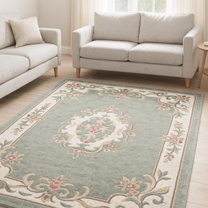 May include: A rectangular area rug featuring a sage green center with a central cream-colored medallion containing pink floral motifs. The border is cream-colored with scrolling green vines and pink flower accents, finished with a thin dark brown edge.