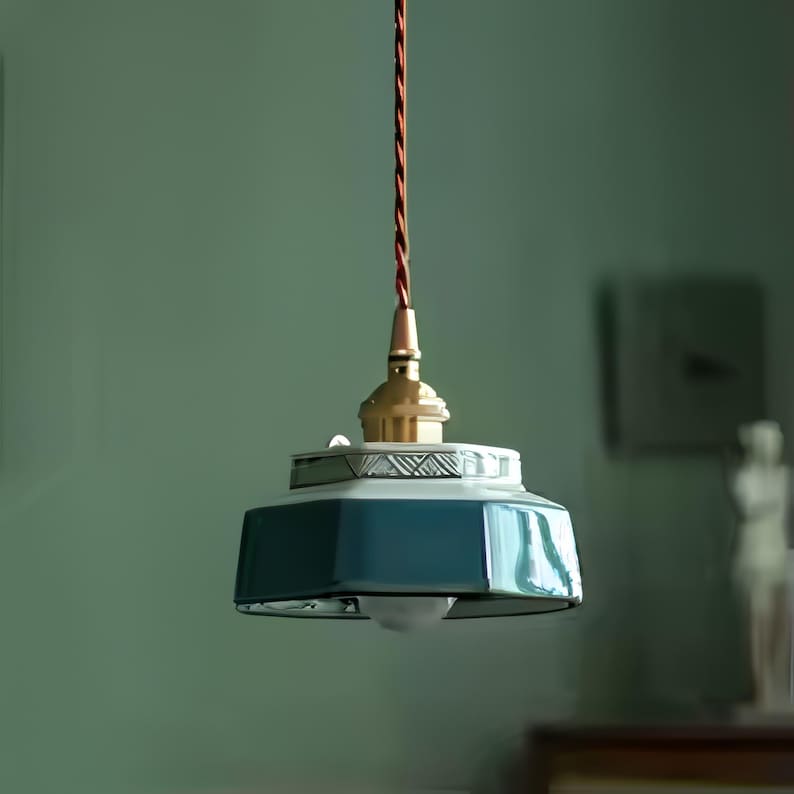 May include: A hanging pendant light with a teal, geometric shade and a brass-colored fixture. The light hangs from a brown, twisted cord. The shade has a white decorative trim. The light is suspended against a muted green wall.