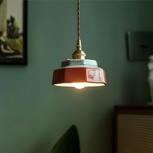 May include: A pendant light fixture with a red, hexagonal shade and a decorative patterned band. The light has a brass-colored fixture and a brown twisted cord. The light is turned on, illuminating the room.