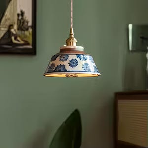 May include: A pendant light fixture with a white and blue floral patterned shade. The light has a brass-colored top and a brown band. The cord is a light brown color.