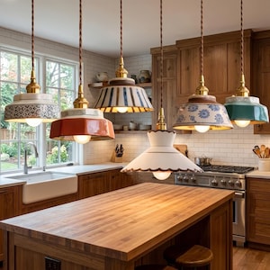 May include: Five pendant lights with ceramic shades hang above a kitchen island. The shades are various colours and patterns, including blue floral, red, and white scalloped edges. The lights have brass fixtures and brown cords.