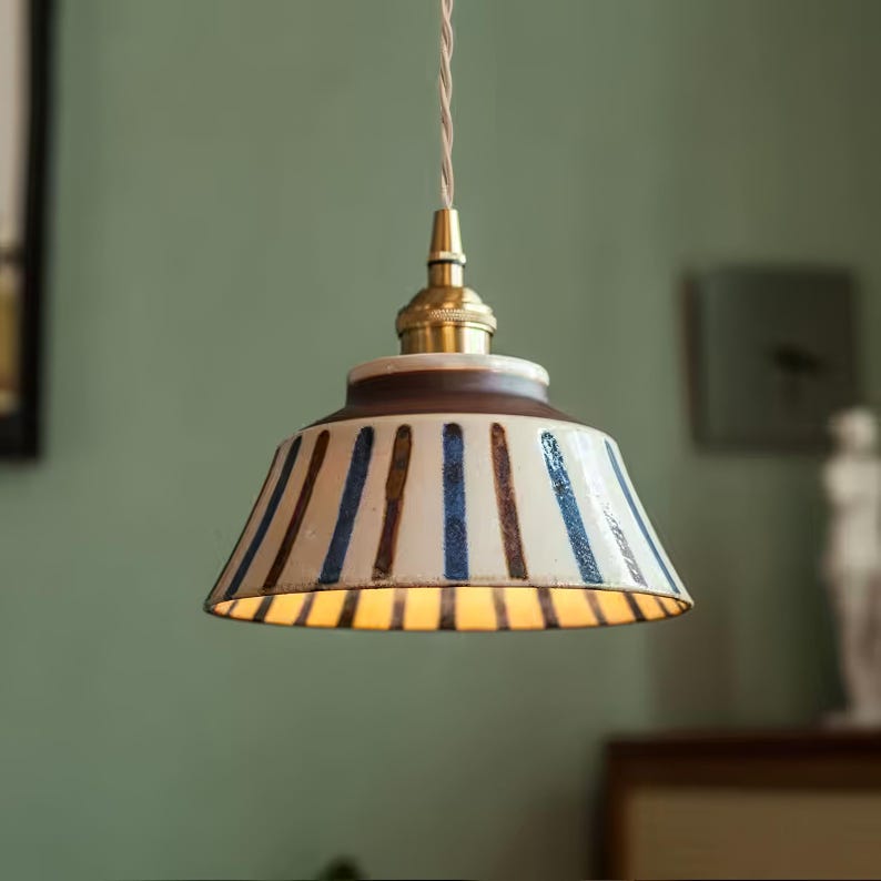 May include: A pendant light fixture with a ceramic shade. The shade is white with vertical blue and brown stripes. The fixture has a brass-colored top and a brown rim. The light is hanging from a white cord.