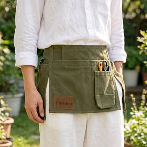 May include: An olive green canvas waist apron featuring a rectangular brown leather patch with the name Christine embossed in black. The apron includes a vertical pocket containing orange and black shears, worn over a white long-sleeve shirt.