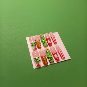 May include: A set of ten artificial nails with a Christmas theme. Designs include a green Grinch, snowflakes, and candy cane stripes. The nails have a clear base with a pink tip against a green background.