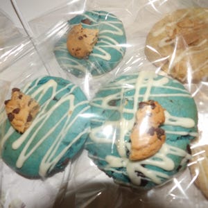 May include: Close-up of several cookies in clear packaging. Some cookies are light blue with white drizzle and a chocolate chip cookie on top. Other cookies are a light brown color with white drizzle.