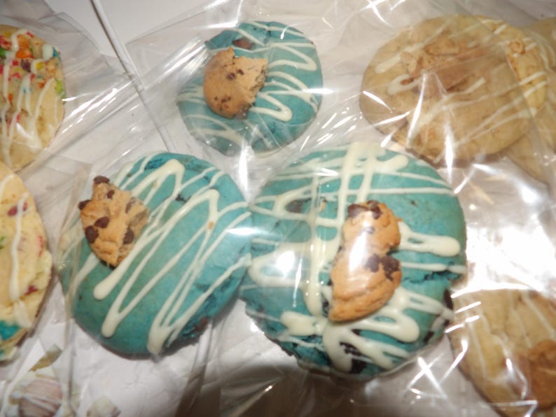 May include: Close-up of several cookies in clear packaging. Some cookies are light blue with white drizzle and a chocolate chip cookie on top. Other cookies are a light brown color with white drizzle.