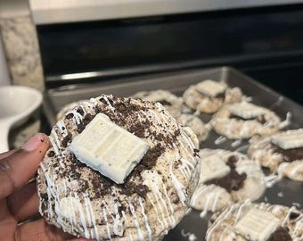 May include: A close-up of a homemade cookie topped with a white chocolate bar. The cookie is covered in crushed biscuits and drizzled with white icing. Other cookies are on a baking tray in the background.