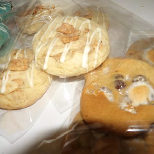 May include: Close-up of several cookies in clear plastic packaging. The cookies are various sizes and shapes, with different toppings. Some cookies have white icing drizzle and cereal pieces. Others have chocolate chips and marshmallows.