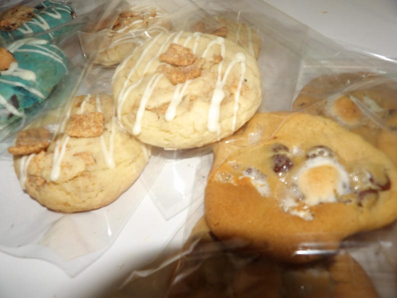 May include: Close-up of several cookies in clear plastic packaging. The cookies are various sizes and shapes, with different toppings. Some cookies have white icing drizzle and cereal pieces. Others have chocolate chips and marshmallows.