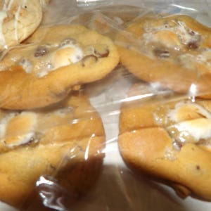 May include: Several golden-brown cookies with melted marshmallows and chocolate chips are visible through a clear plastic bag. One cookie with white icing is also visible. The cookies appear soft and freshly baked, perfect for a sweet treat.