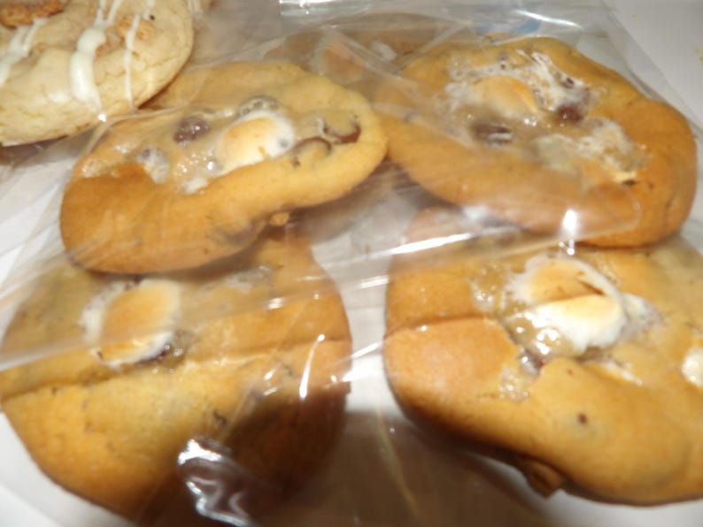 May include: Several golden-brown cookies with melted marshmallows and chocolate chips are visible through a clear plastic bag. One cookie with white icing is also visible. The cookies appear soft and freshly baked, perfect for a sweet treat.